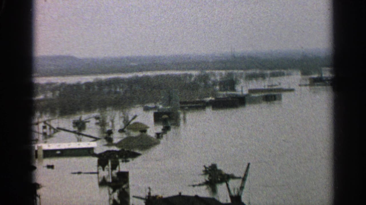 1965 Minnesota 8mm view of a flooded waterside work area, with platforms, crane-like equipment, and low hills across the water.