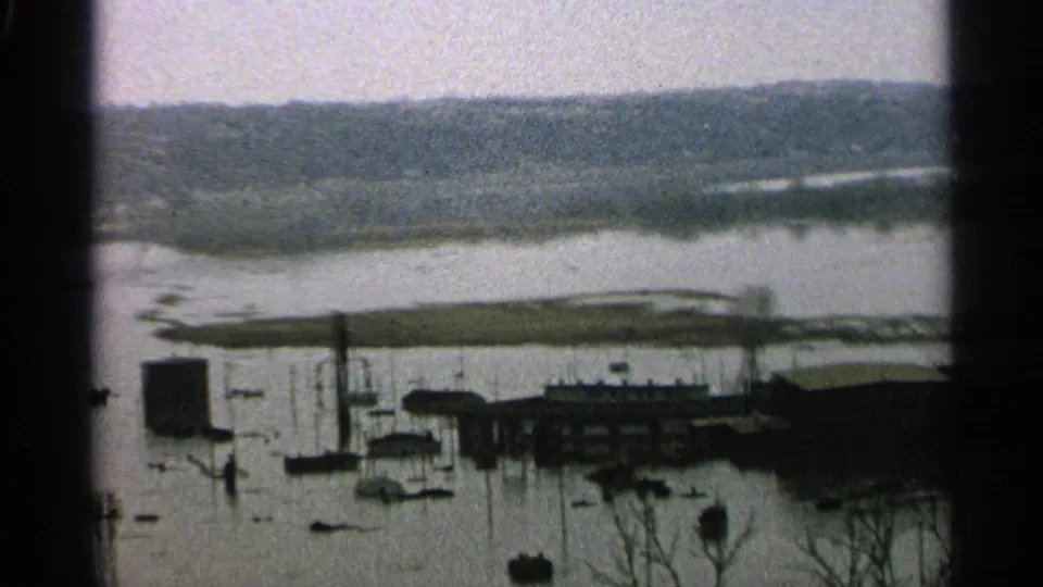 1965 Minnesota 8mm view of a flooded lakeside waterfront, with docks, small boats, low buildings, bare branches, and distant shoreline.