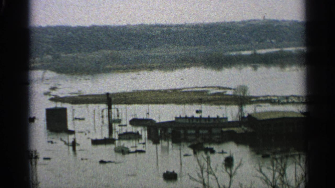 A flooded Minnesota lakeside marina or harbor area appears in 8mm footage, with boats, dock structures, poles, buildings, and wooded shore beyond.