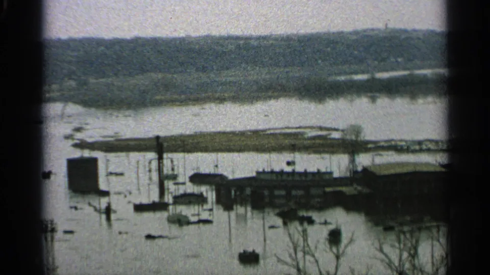 A flooded Minnesota lakeside marina or harbor area appears in 8mm footage, with boats, dock structures, poles, buildings, and wooded shore beyond.