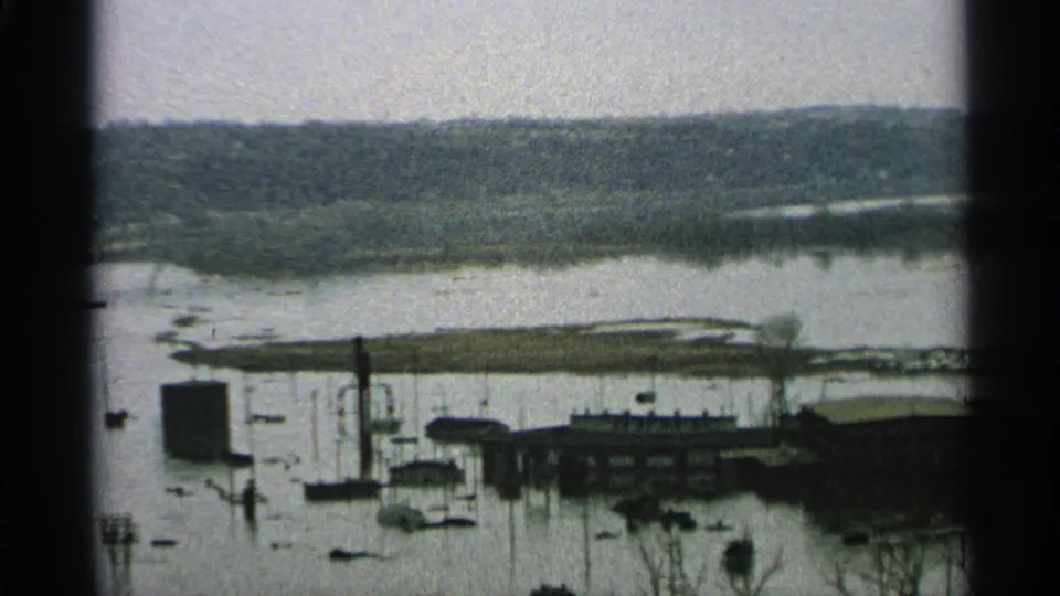 1965 8mm footage shows floodwater across a lakeside community area, with submerged dock lines, boats, waterfront buildings, and bare trees in front.