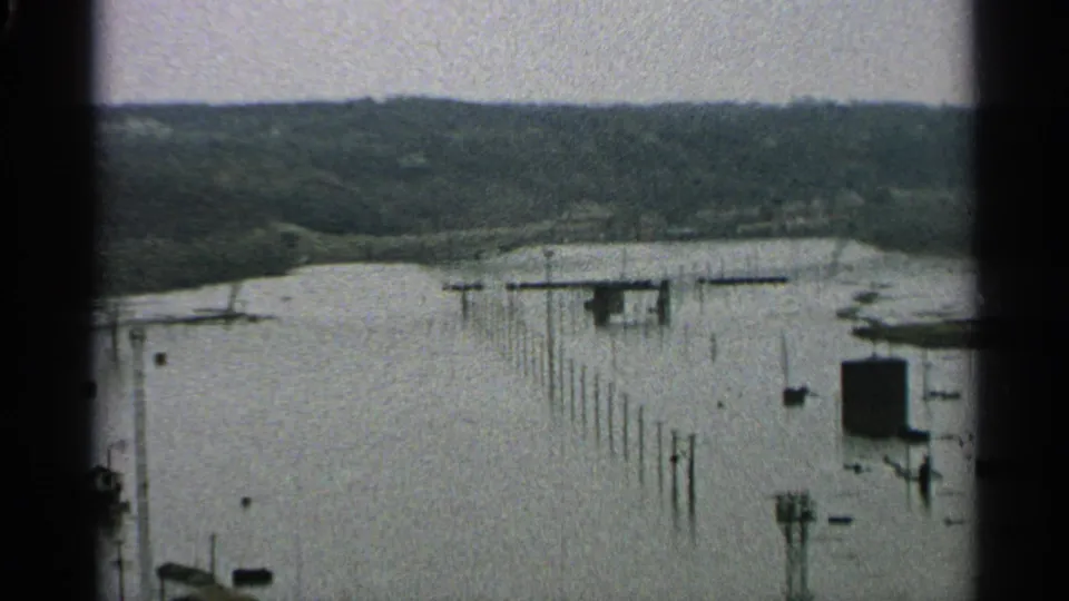 The final 1965 Minnesota frame widens across floodwater, showing rows of poles or dock supports, small boats, a building edge, and a wooded shoreline.