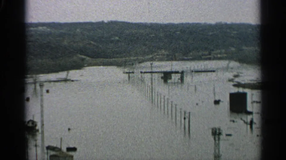 1965 Minnesota 8mm frame showing high water around dock posts, small structures, and a wooded hillside beyond the river.