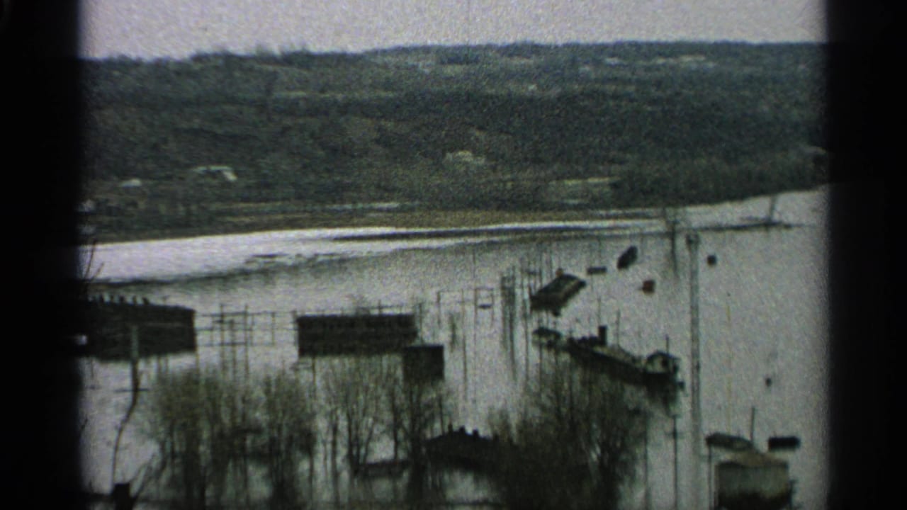 Flooded waterfront with docks, small buildings, bare trees, and hills.