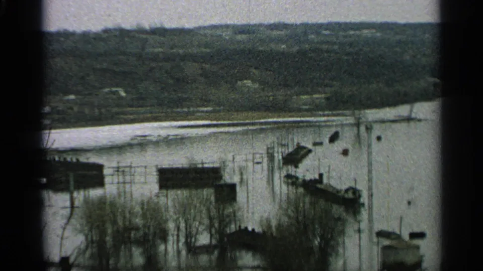 1965 Minnesota 8mm frame of a flooded riverside area with dock structures, bare trees, small buildings, and wooded hills in the distance.