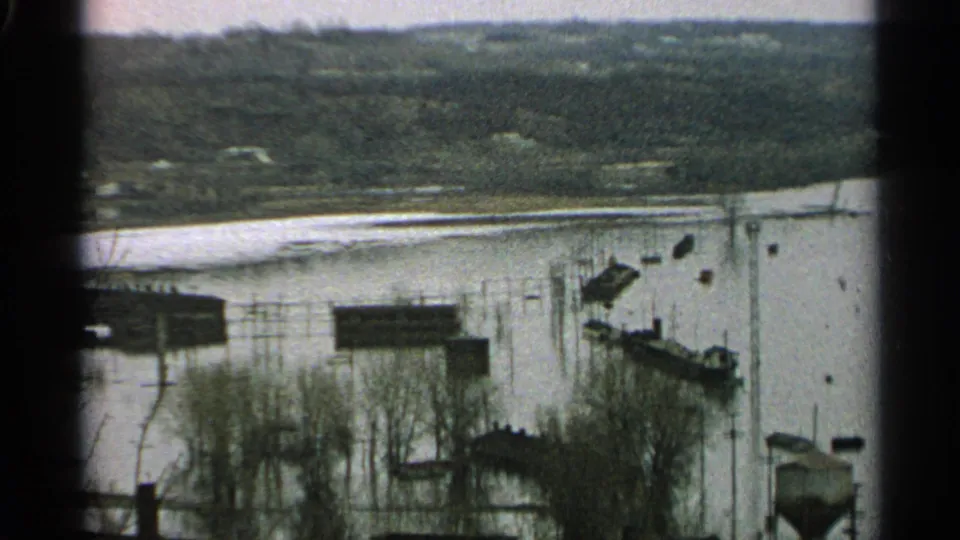1965 Minnesota home-movie frame showing high water around riverside buildings, dock sections, bare trees, and a wooded background.