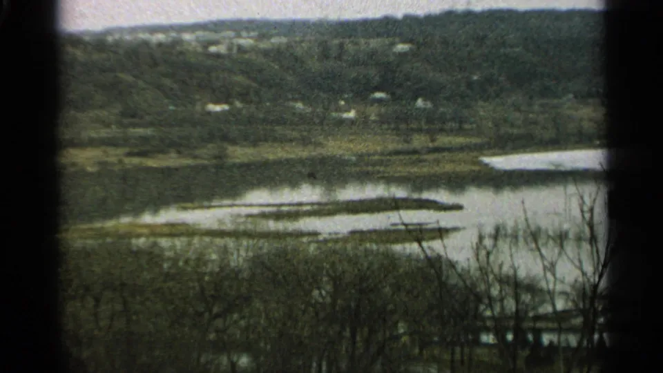1965 Minnesota 8mm frame showing calm water, a wooded far shore, scattered houses, and dark foreground branches.