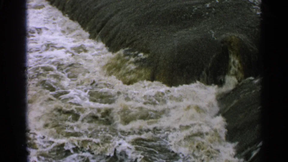 1965 Minnesota home-movie frame showing white water moving through a dark concrete spillway or dam channel.