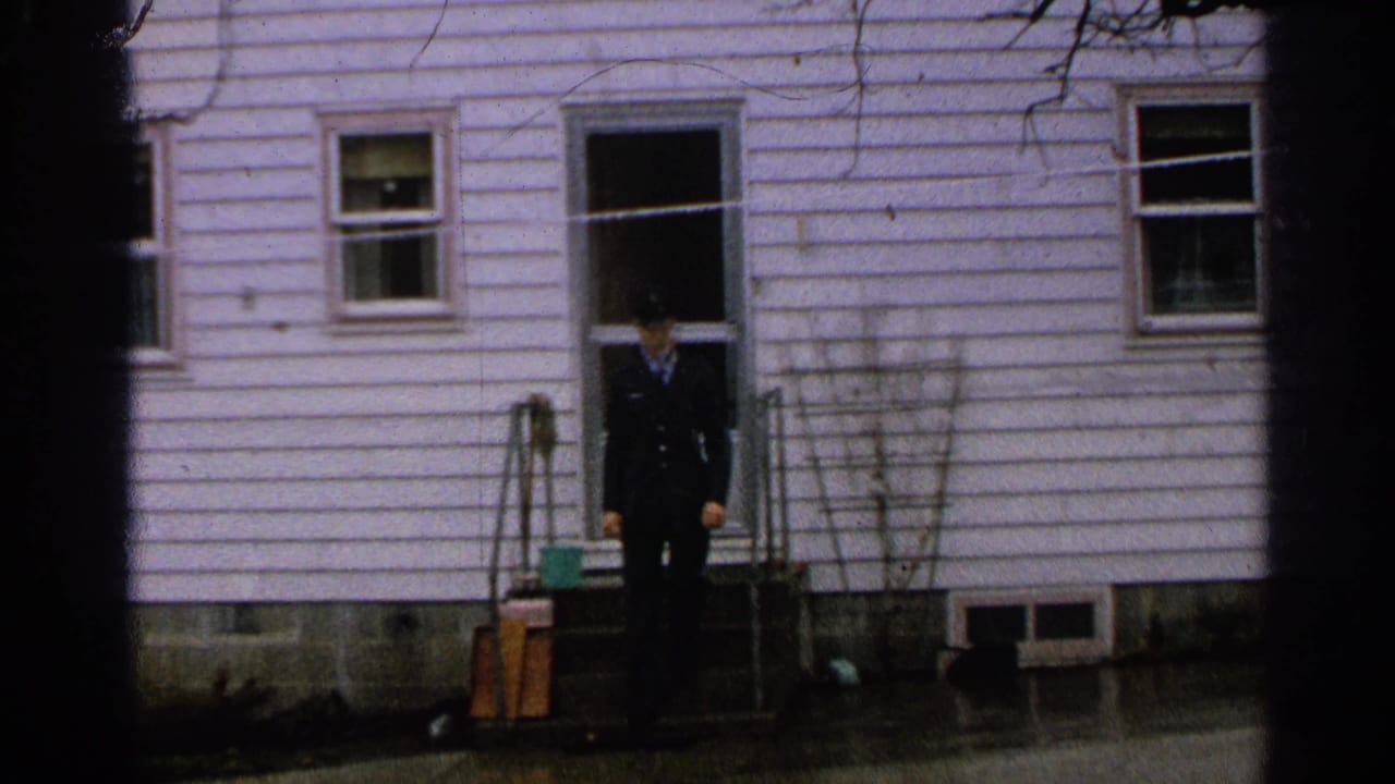 In 1965 Minnesota footage, a man in a dark uniform stands at an open doorway of a white-sided house with wet pavement below.