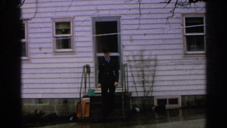 In 1965 Minnesota footage, a man in a dark uniform stands at an open doorway of a white-sided house with wet pavement below.