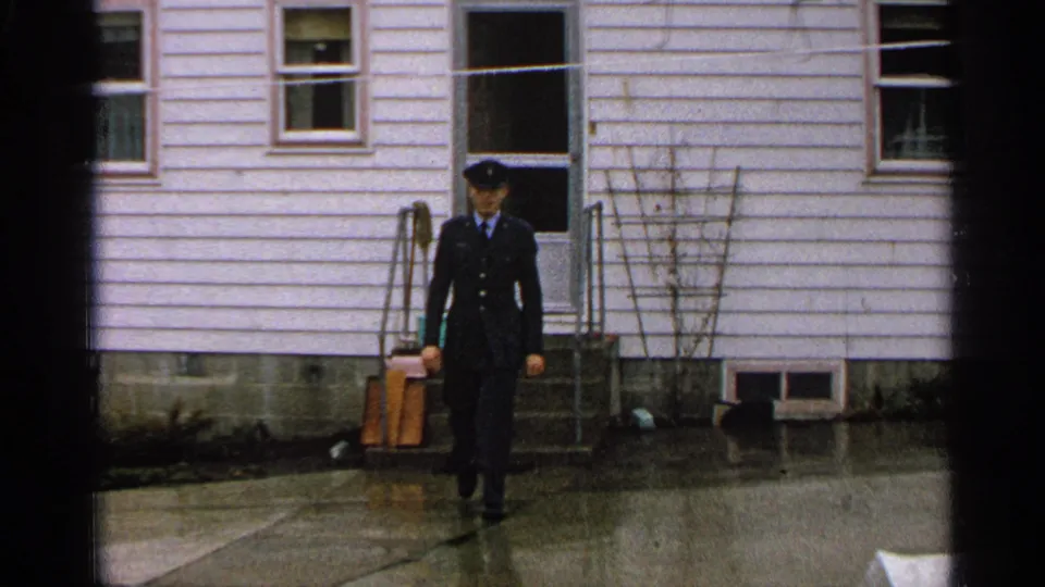 The 1965 Minnesota home-movie frame shows the uniformed man walking away from the white house across rain-wet pavement.
