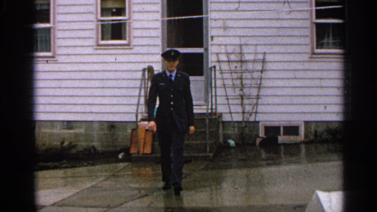 In 1965 Minnesota home-movie footage, a young man in a dark uniform and cap walks toward the camera outside a white-sided house.