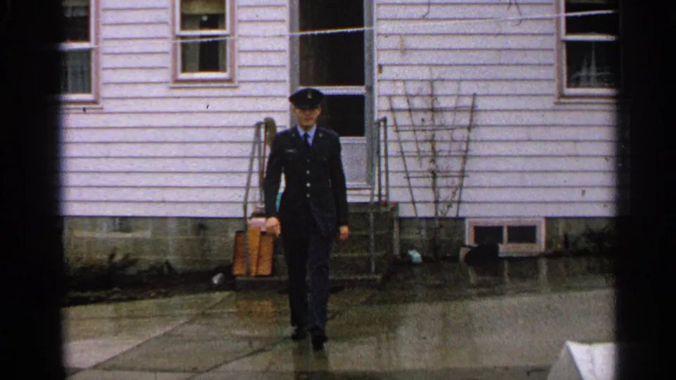 In 1965 Minnesota home-movie footage, a young man in a dark uniform and cap walks toward the camera outside a white-sided house.