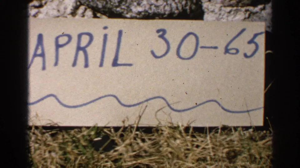 The 8mm frame shows the handwritten “APRIL 30-65” card in close view, with grass and film borders visible.