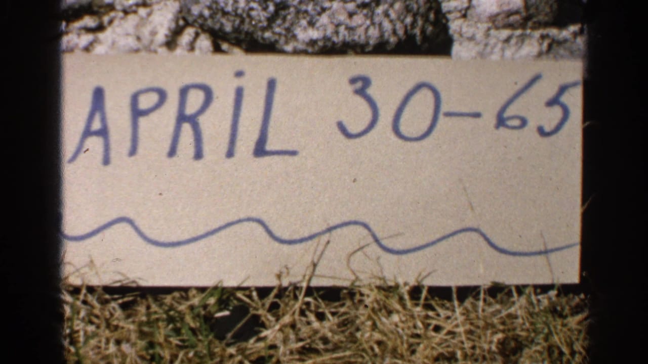 Handwritten April 30-65 card propped on a rock above dry grass.
