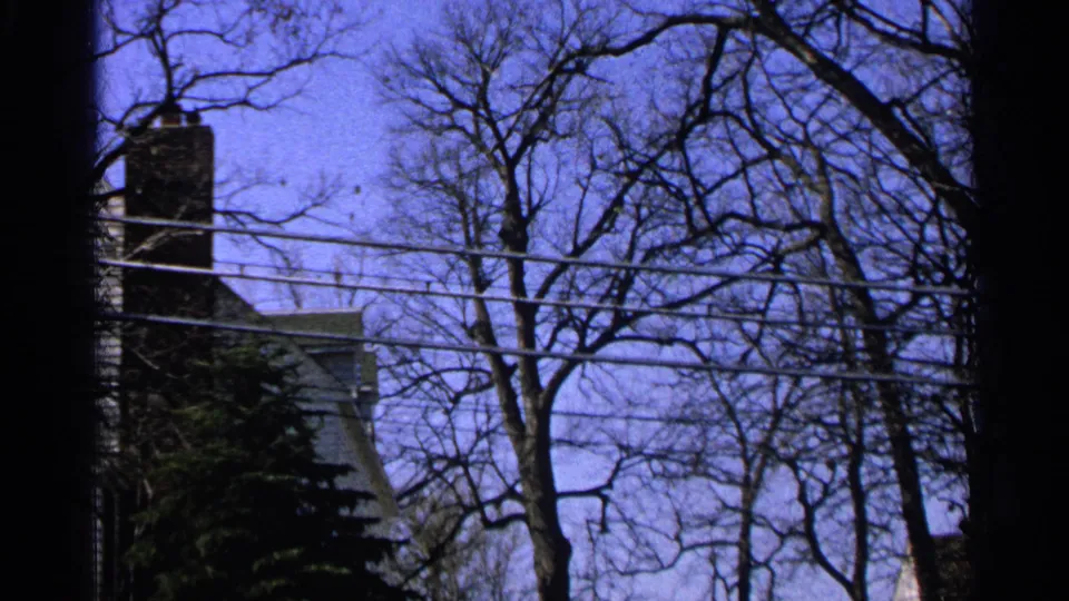 A grainy 8mm view of a house corner with brick chimney, overhead wires, and leafless trees in Minnesota, 1965.