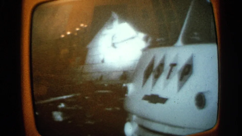 1965 Minnesota home-movie frame showing a closer televised view of a damaged car next to a van with large lettering.