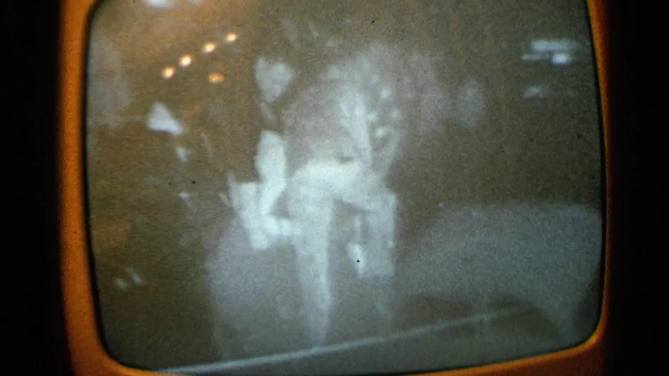 1965 Minnesota home-movie frame of a grainy TV broadcast with a faint person visible near indistinct vehicles or debris.