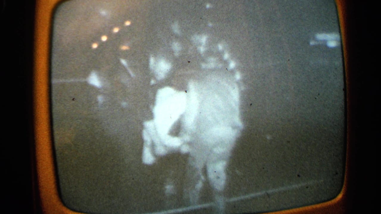 1965 Minnesota 8mm frame of a rounded TV screen with blurred monochrome footage of two indistinct figures, possibly in an emergency scene.