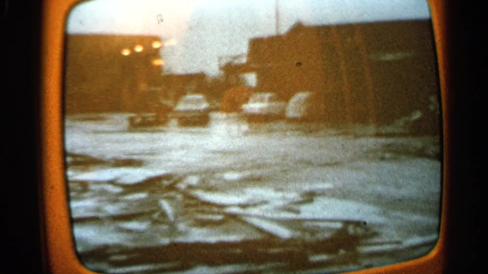 1965 Minnesota 8mm frame of a TV-viewed street or lot with parked mid-century cars, scattered debris, and low buildings.