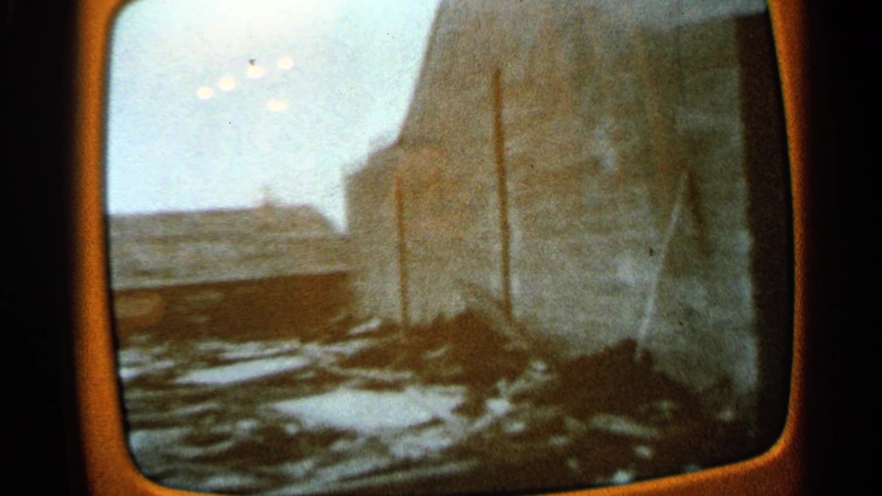 1965 Minnesota 8mm frame showing a damaged rural structure, snow patches, and scattered debris seen through a rounded screen-like border.