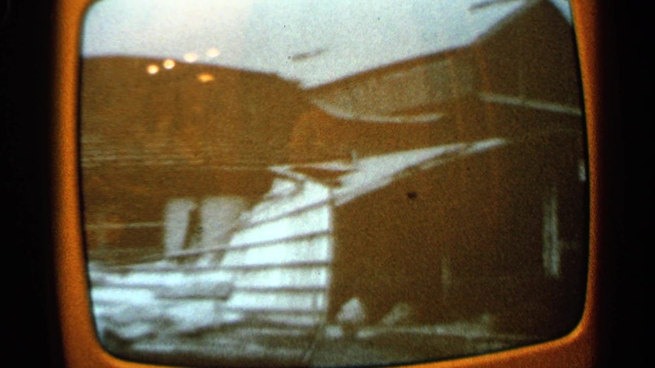 1965 Minnesota 8mm frame with damaged wooden siding, a dark building edge, snow on the ground, and rural structures in the background.