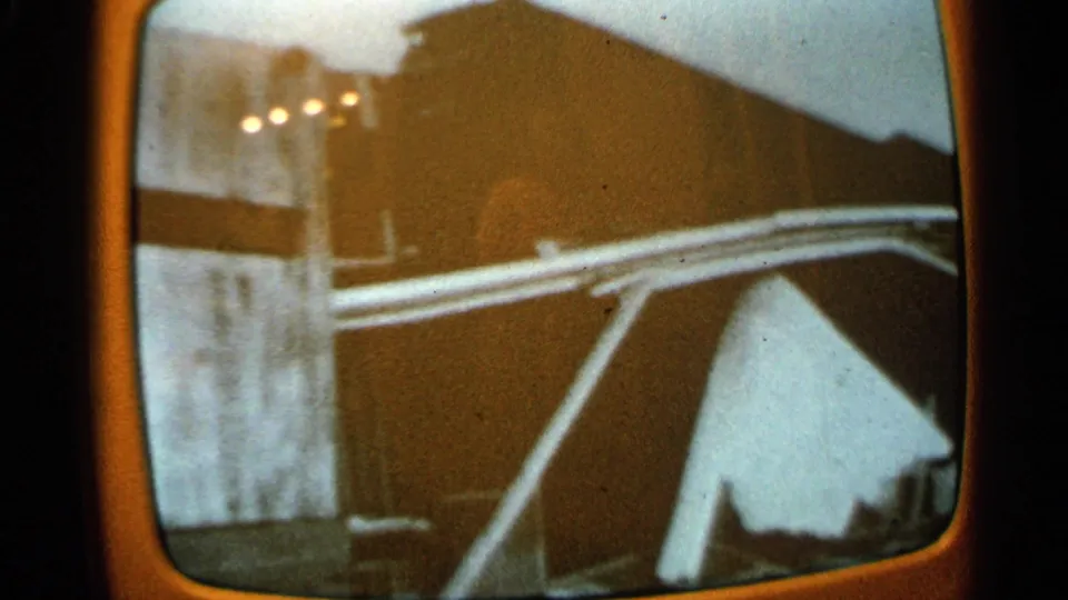 Faded 8mm frame of a television showing a damaged building interior with a dark sloped roof shape and pale support beams.