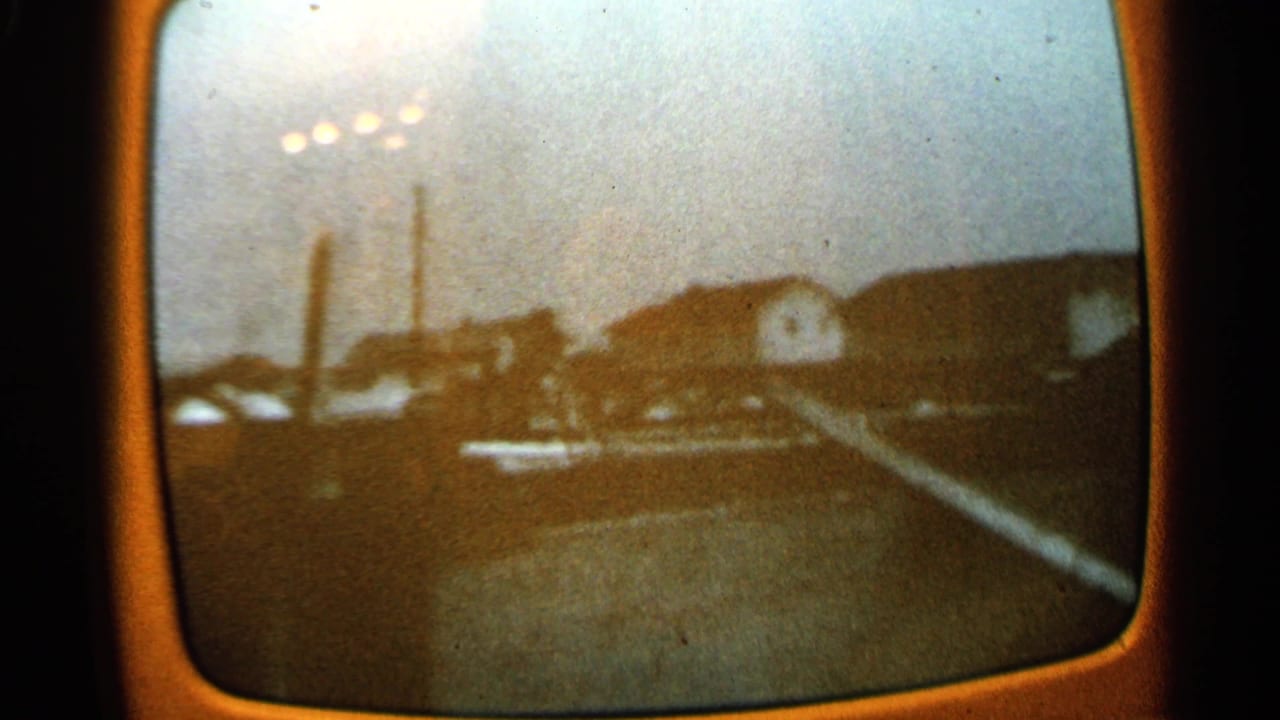 Industrial buildings and stacks behind a dark foreground, viewed through a rounded screen with visible film grain.