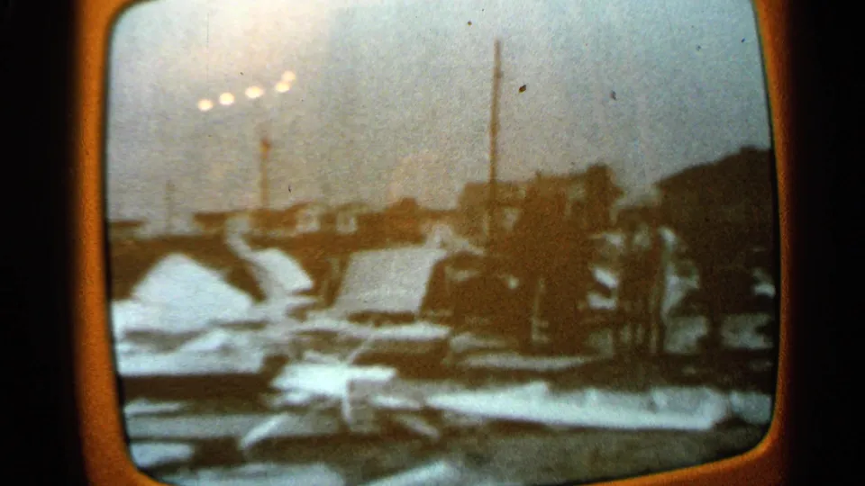 Minnesota 1965 home-movie frame showing a closer look at damaged material or wreckage near industrial buildings, parked cars, and tall stacks.