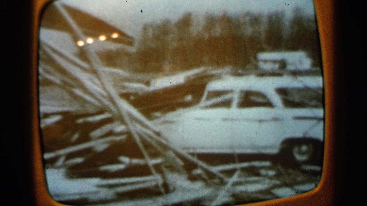 Orange television screen showing a white station wagon amid snow, fallen branches, and debris.