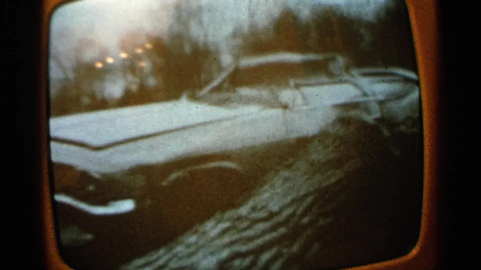 1965 Minnesota 8mm frame of a television broadcast showing a white station wagon by snowy storm debris and bare trees.