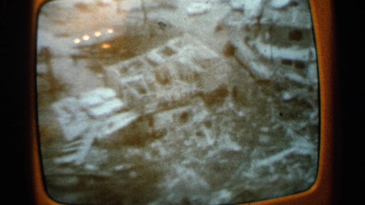 Rounded television screen showing grainy aerial flood damage and scattered structures.
