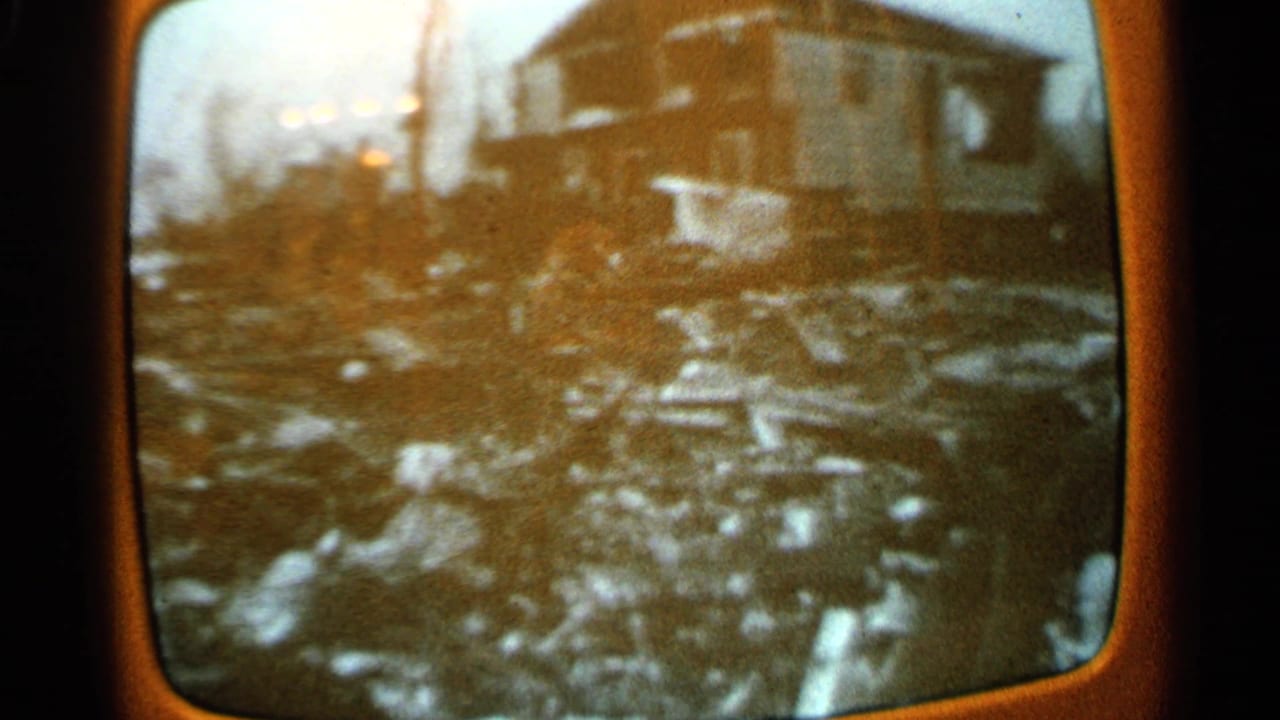 Television screen showing a damaged house and debris-strewn yard.