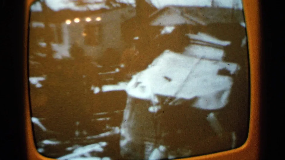 Wider 8mm frame of the orange television screen showing debris, bright points of light, and a damaged white vehicle or panel.