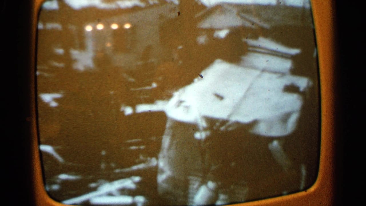 Person standing amid debris near a damaged white vehicle or structure in a grainy 8mm frame.