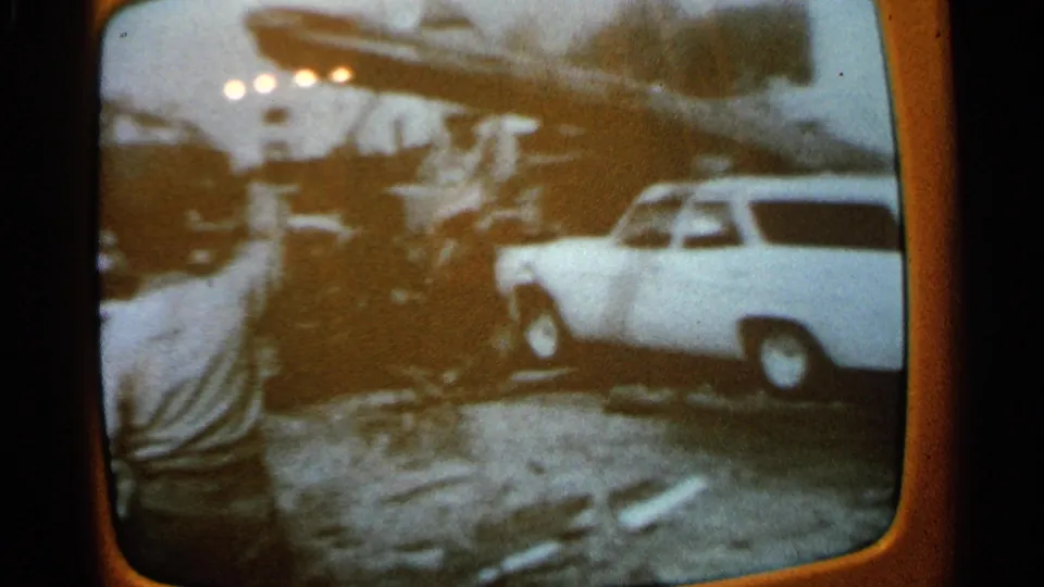 1965 Minnesota 8mm frame showing a white station wagon near a large fallen branch and scattered storm debris.