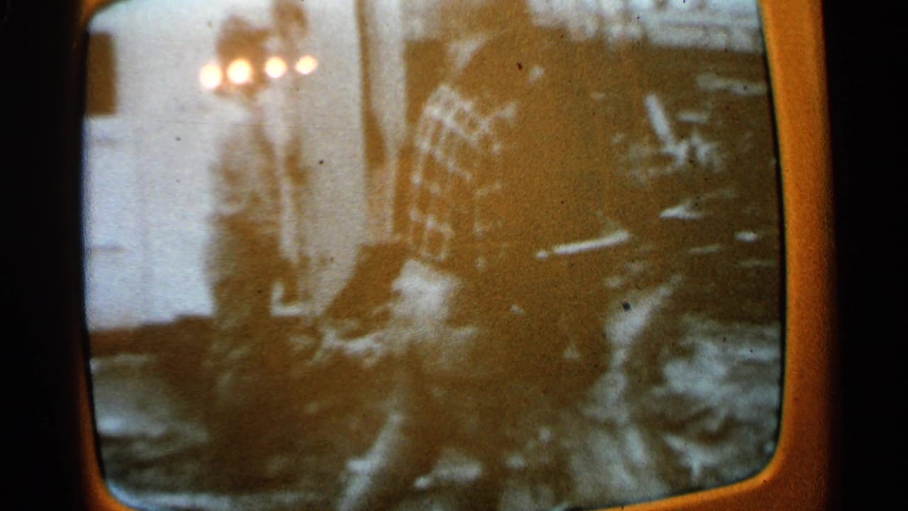 1965 Minnesota 8mm frame of a vintage television screen with indistinct figures, room reflections, and warm orange screen edges.