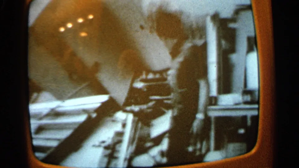 Close 8mm view of a vintage TV broadcast, with a blurred figure leaning near equipment or debris and reflections on the glass.