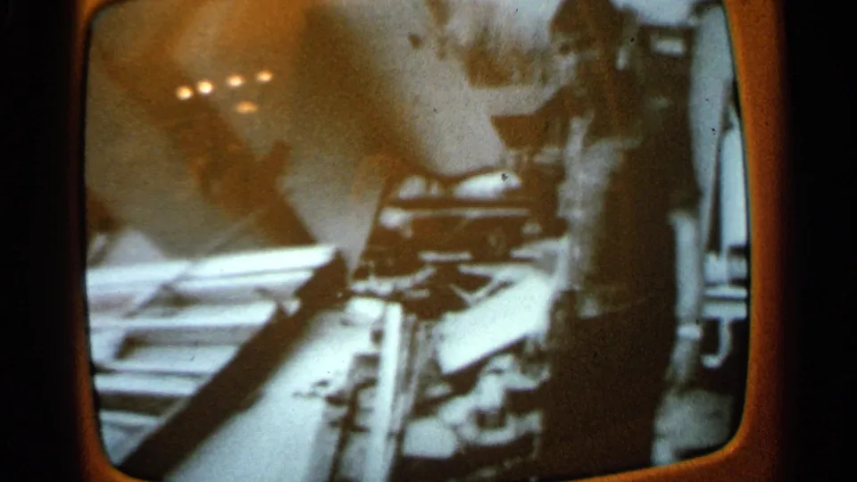 1965 home-movie frame of a television screen showing a blurred figure and angular debris-like objects under warm interior reflections.