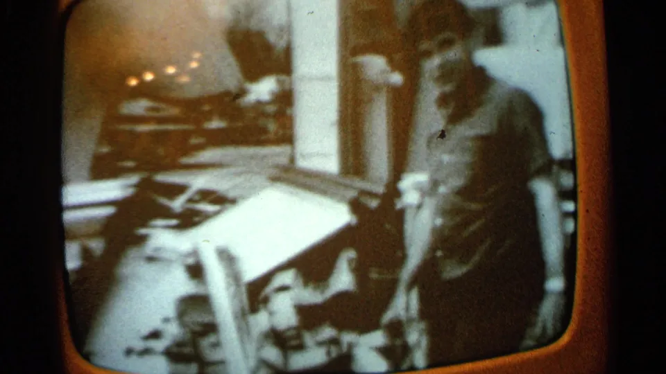 The man appears to gesture or point near outdoor objects, with a parked car and entry area behind him.