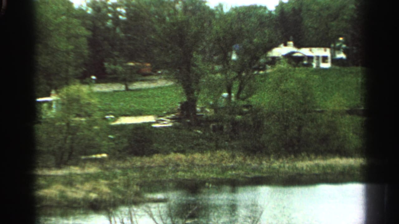 1965 Minnesota 8mm frame showing a stream, grassy slope, trees, and a distant light-colored house on a hill.