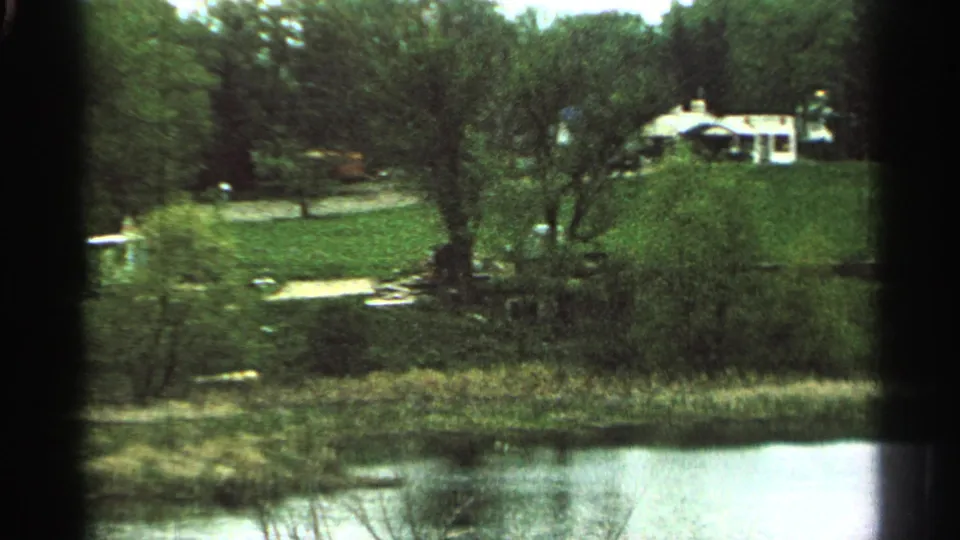 1965 Minnesota 8mm frame showing a stream, grassy slope, trees, and a distant light-colored house on a hill.