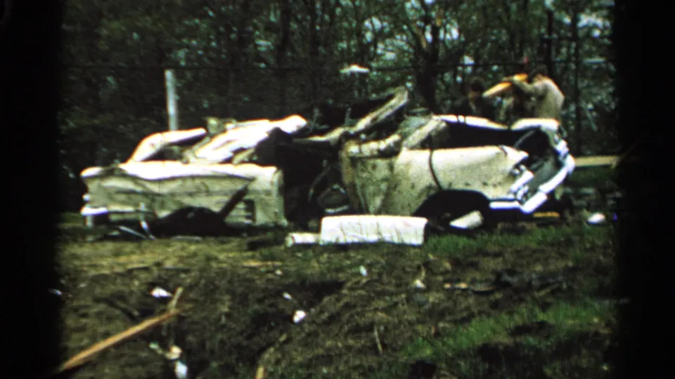 1965 Minnesota 8mm frame showing a badly crushed white and blue car body on muddy grass with debris and one person near the wreck.