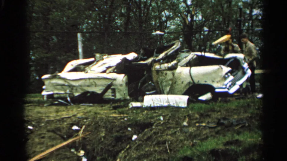 1965 Minnesota 8mm frame of a crushed white and blue automobile in a wooded area, with two people visible near the wreckage.