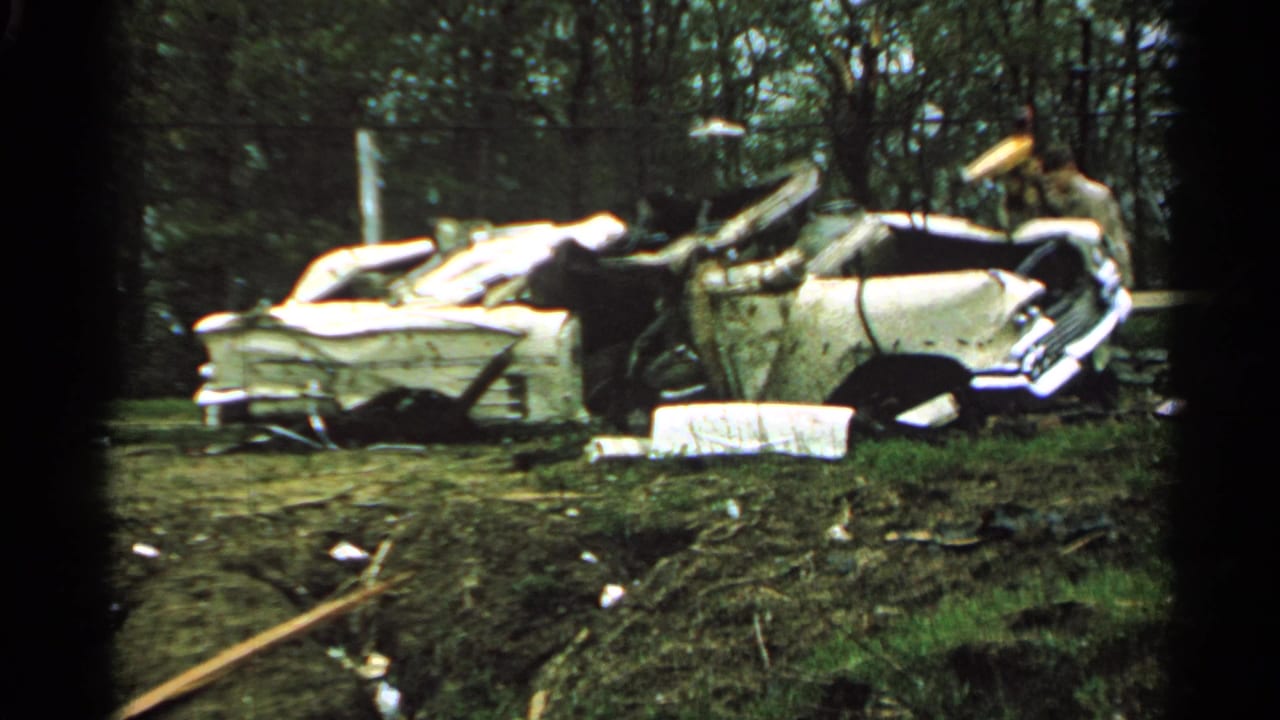 Crushed white car in a debris-strewn grassy area after storm damage.