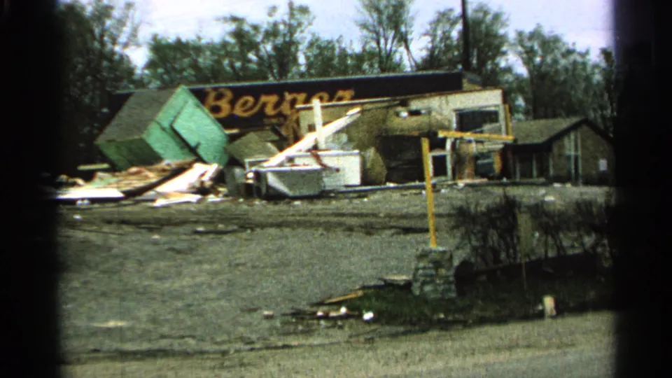 1965 Minnesota 8mm frame showing roadside storm debris, a damaged building, and the partial Berger sign in the background.