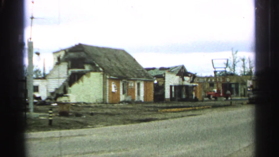 1965 Minnesota 8mm frame showing a street lined with damaged small buildings and exposed roof areas under an overcast sky.