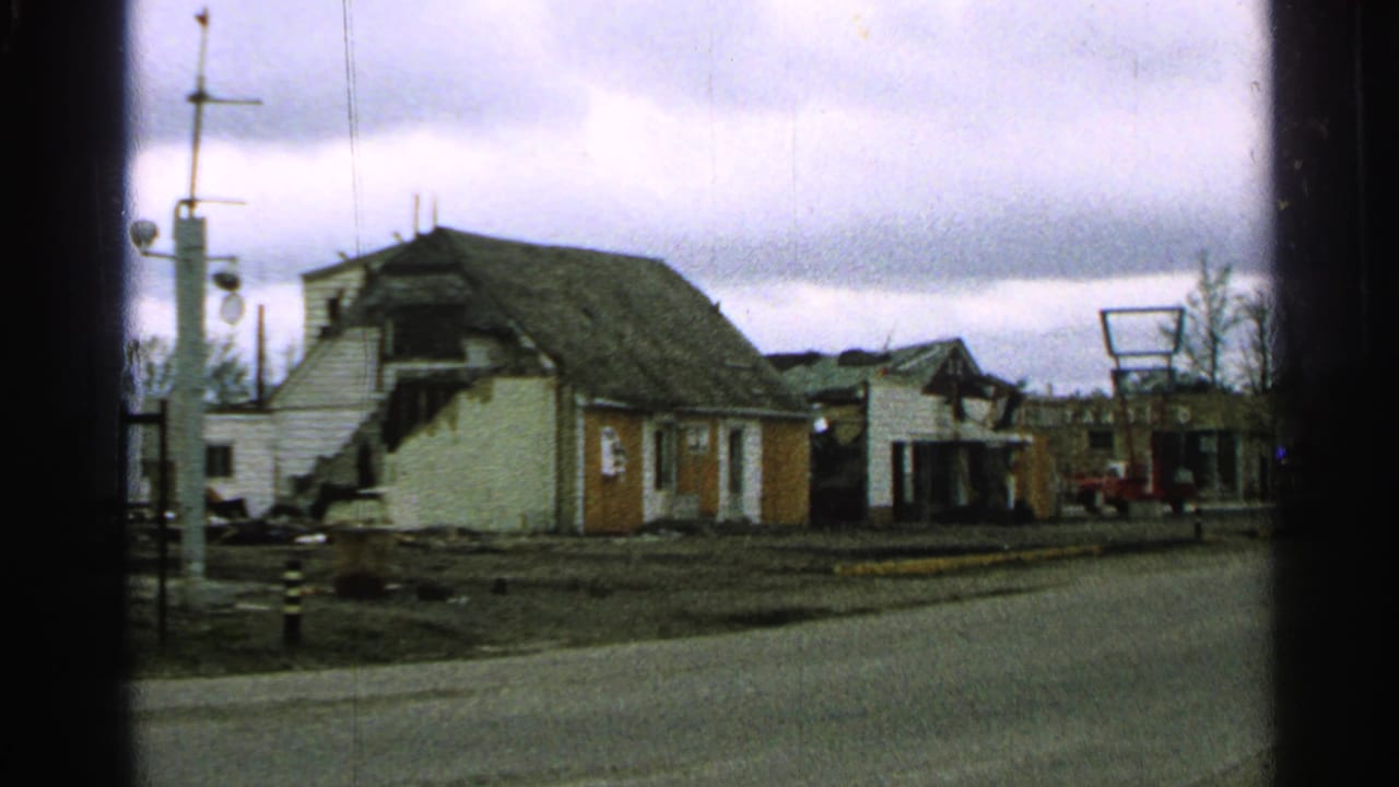 1965 Minnesota home-movie frame showing a damaged house, utility poles, and a quiet street under gray clouds.