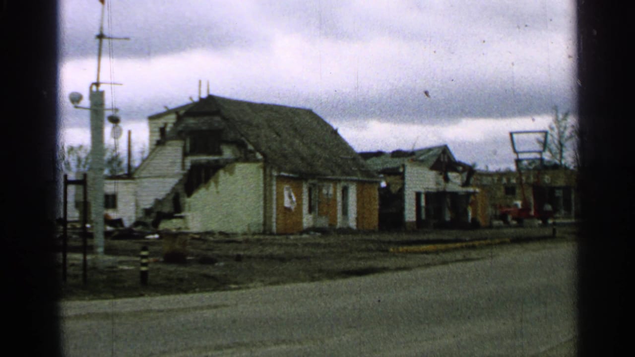 1965 Minnesota exterior with a damaged house, sign frame, utility poles, and a red truck in the distance.
