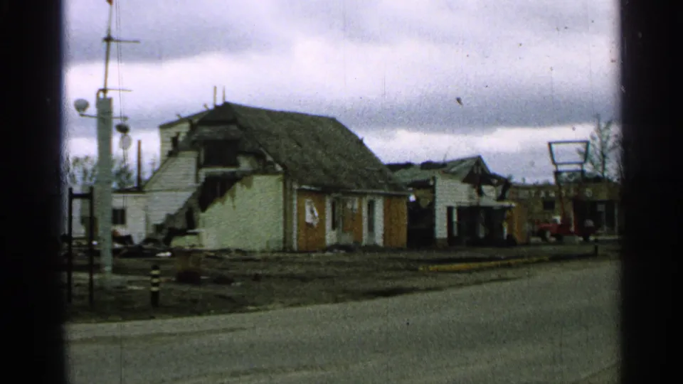 1965 Minnesota exterior with a damaged house, sign frame, utility poles, and a red truck in the distance.